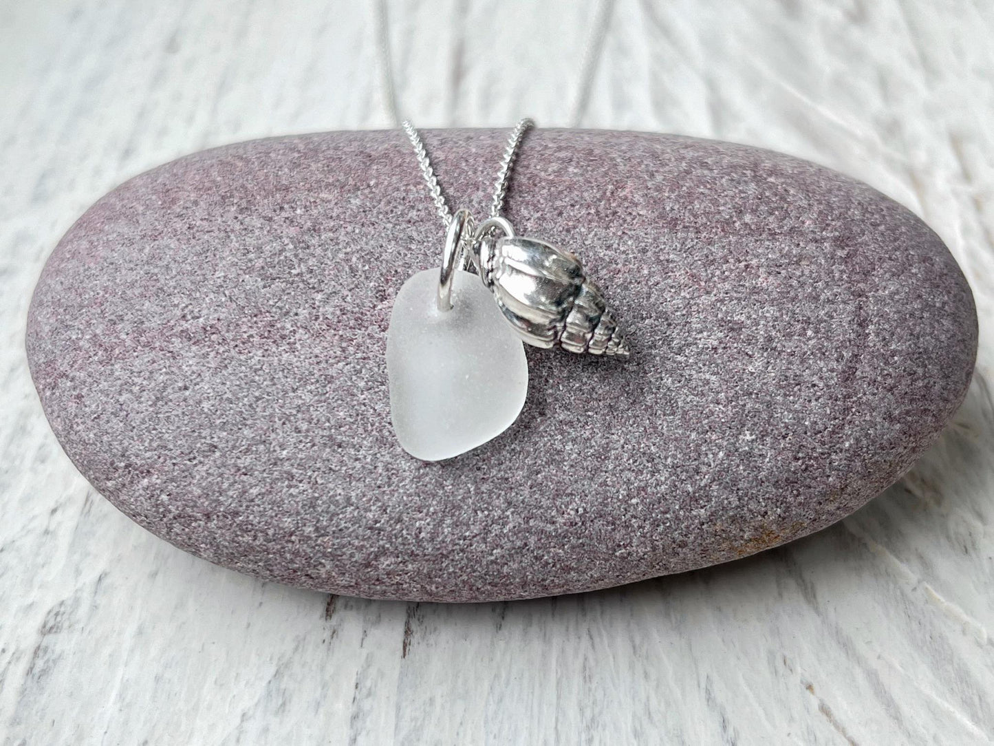 Necklace with white sea glass and silver shell pendant laid on a purple pebble against a textured surface