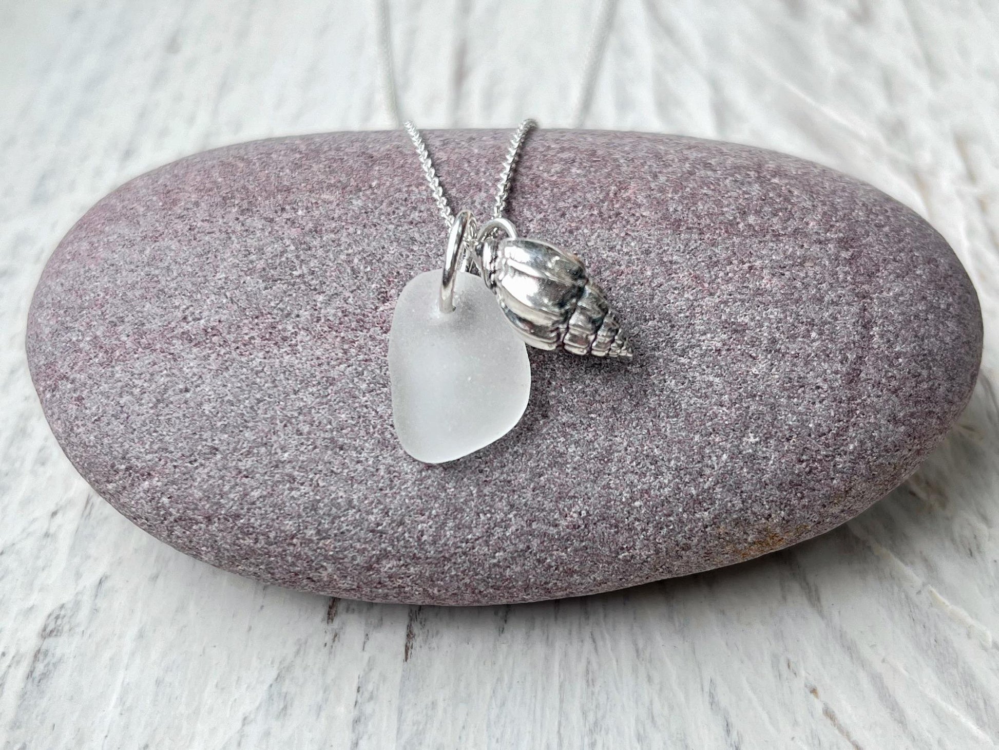 Necklace with white sea glass and silver shell pendant laid on a purple pebble against a textured surface