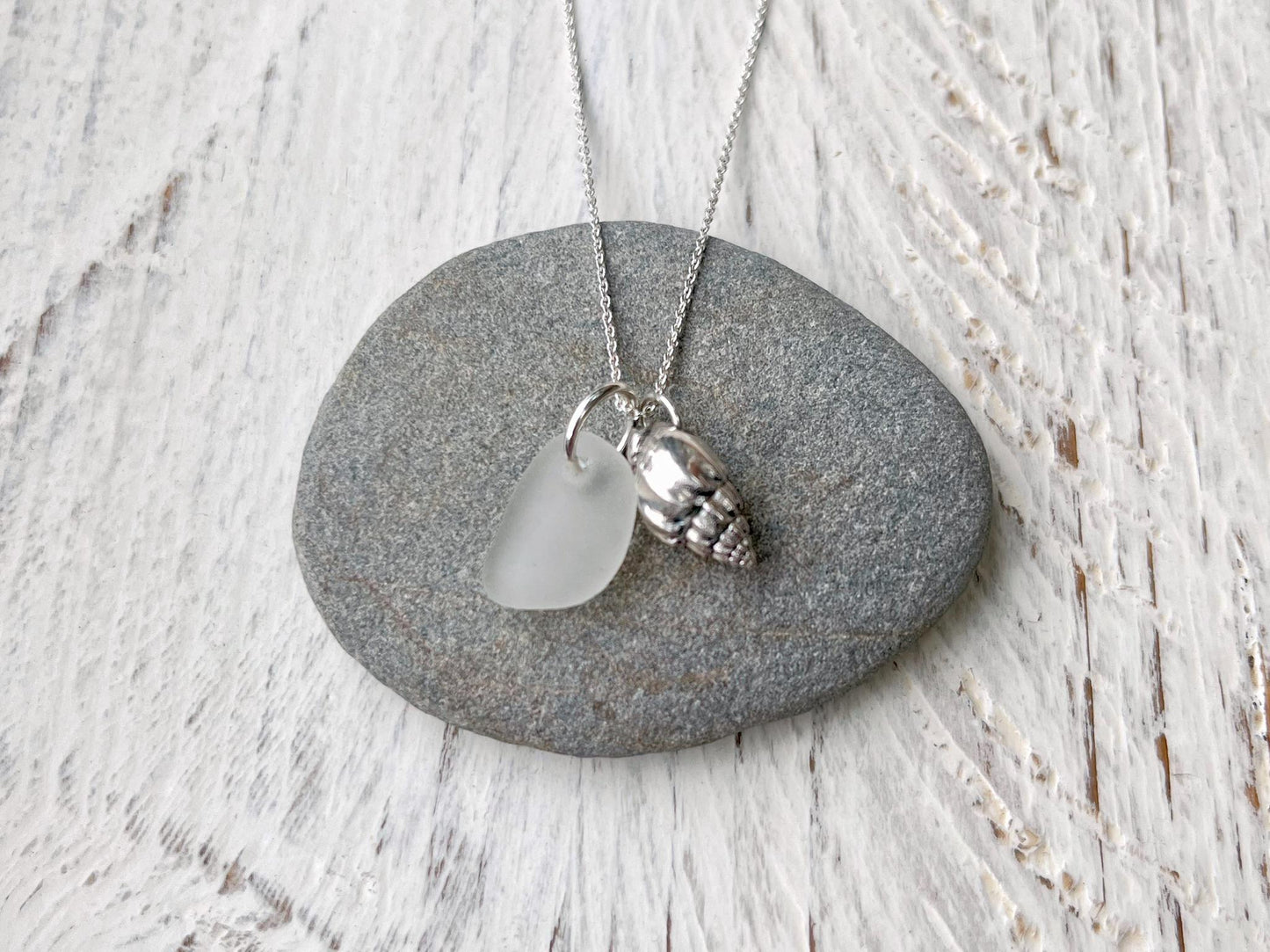 Necklace with white sea glass and silver shell pendant laid on a grey pebble against a textured surface