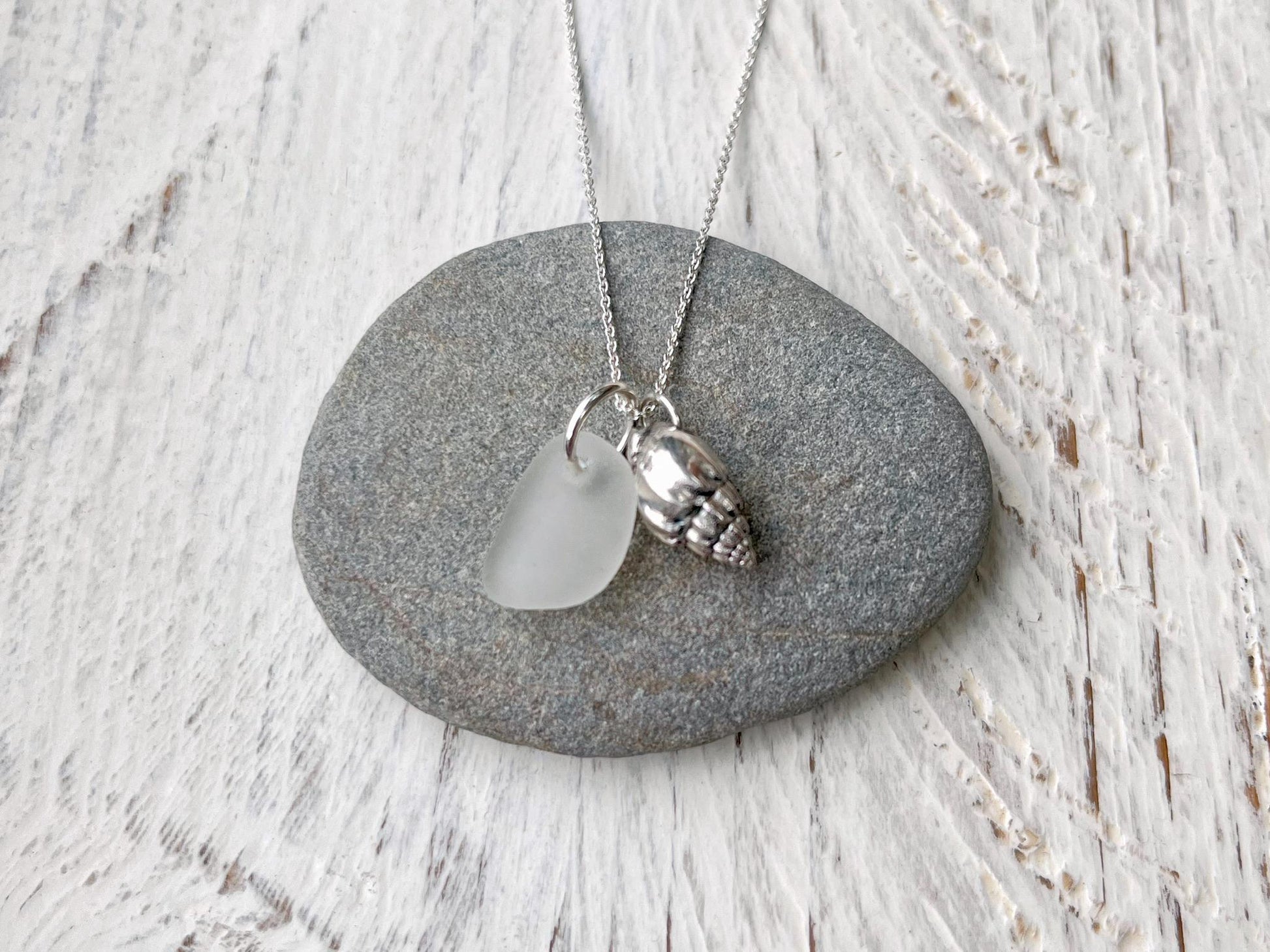 Necklace with white sea glass and silver shell pendant laid on a grey pebble against a textured surface