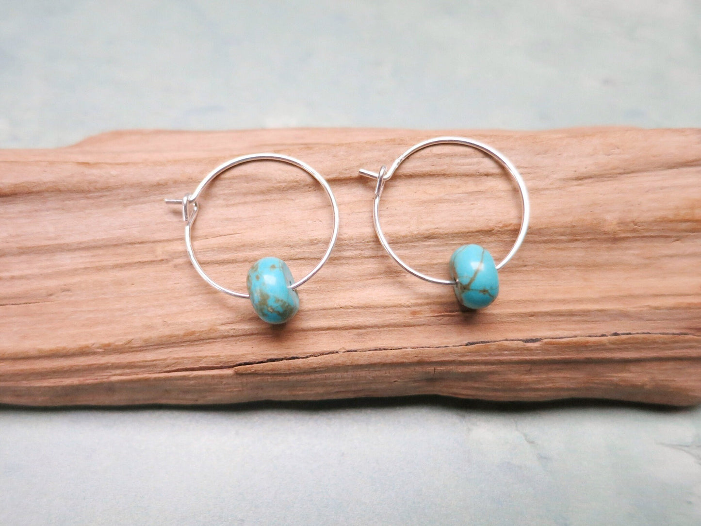 Silver hoop earrings with turquoise beads laid on driftwood against a pale background