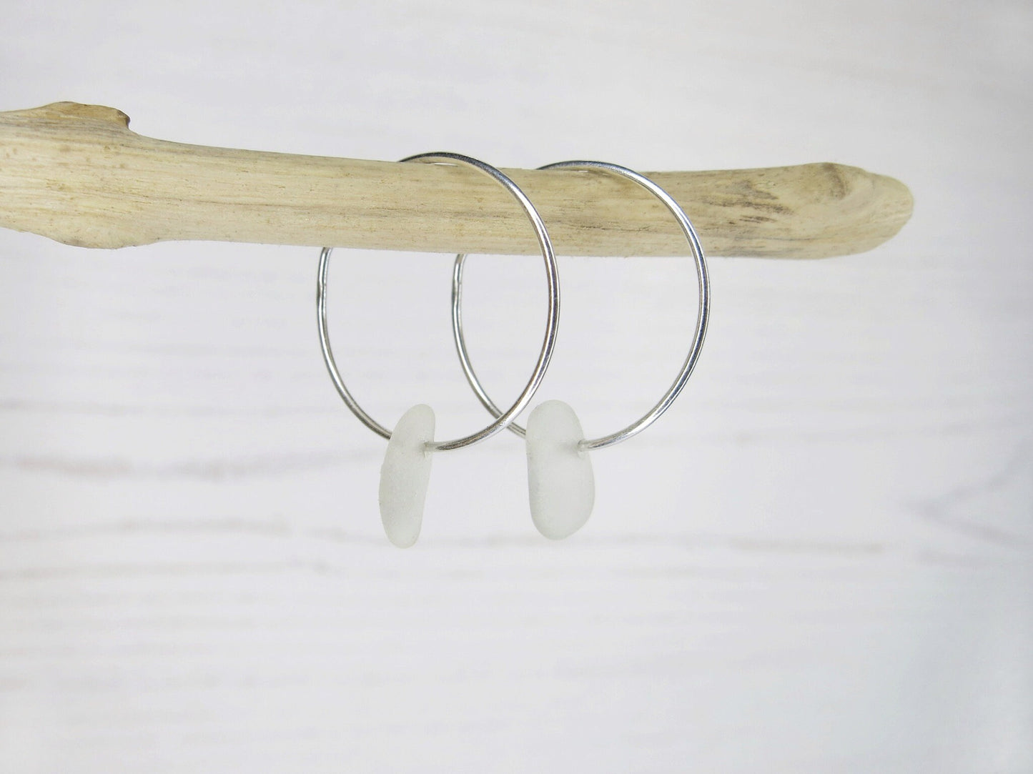 Silver hoop earrings with white sea glass hanging on driftwood against a white background