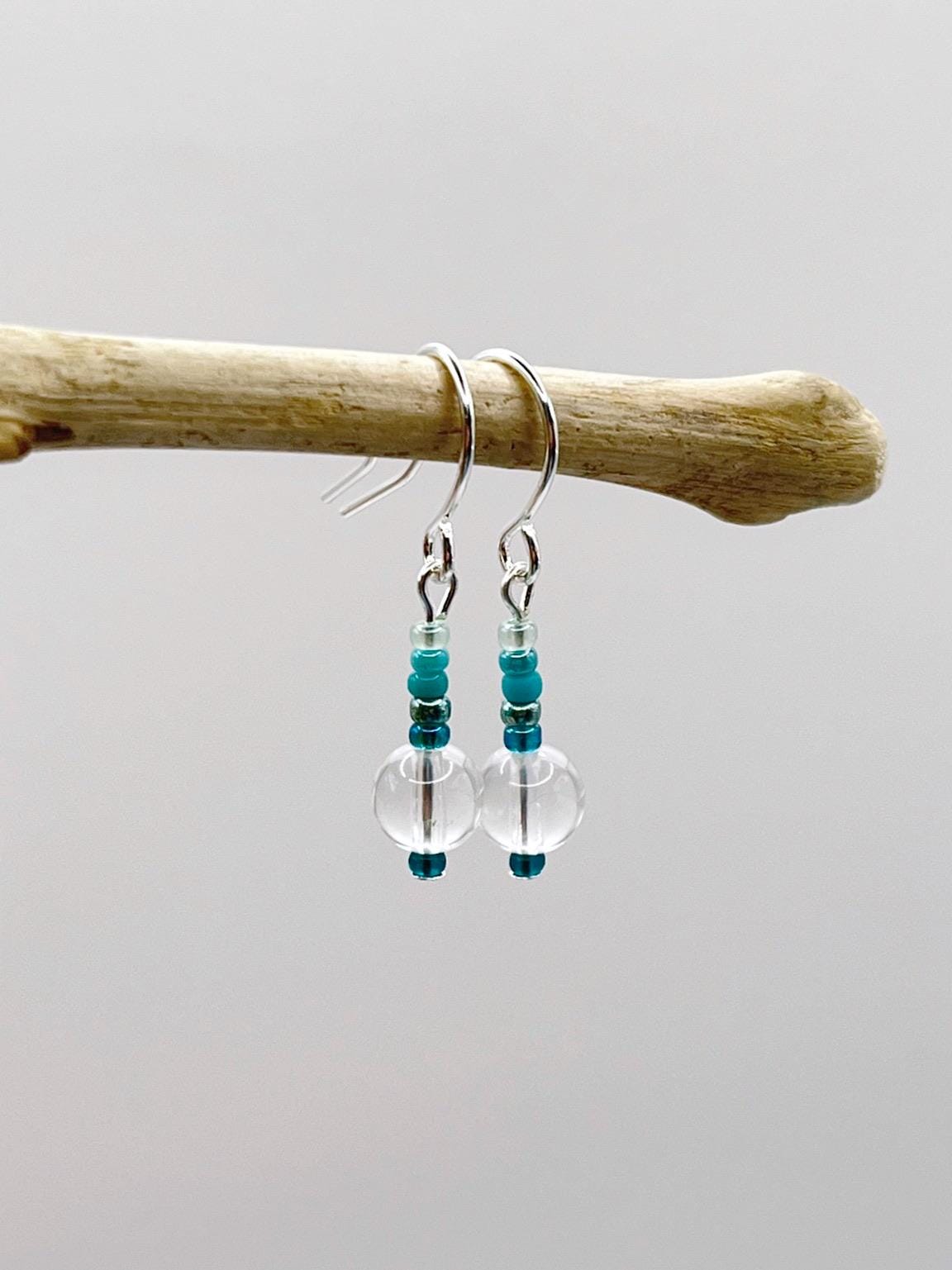 Pair of earrings with clear crystal and turquoise seed beads hanging on driftwood against a white background