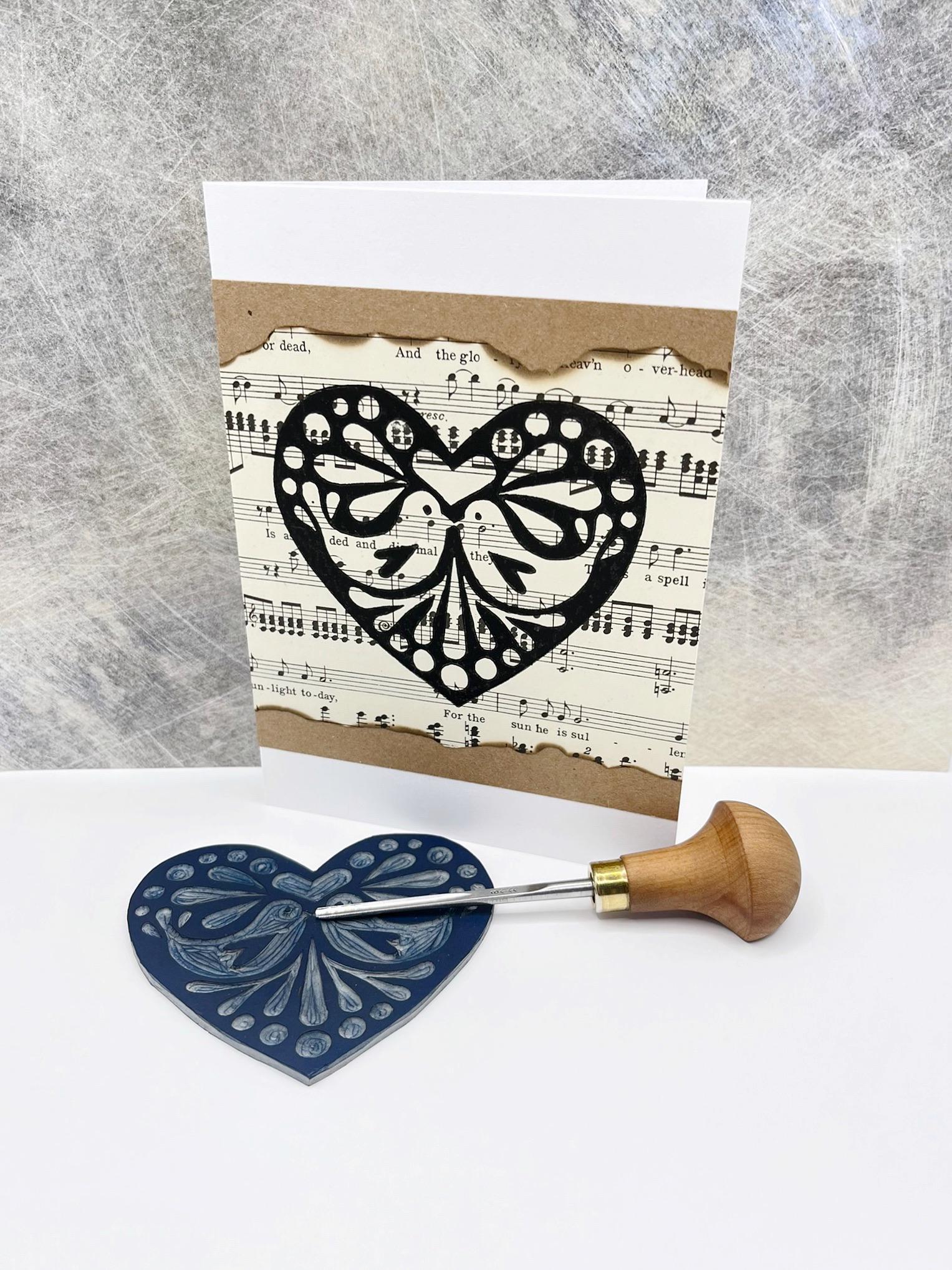 Card with an original lino printed black heart design on sheet music. Photo includes lino and cutting tool used stood against a textured background.