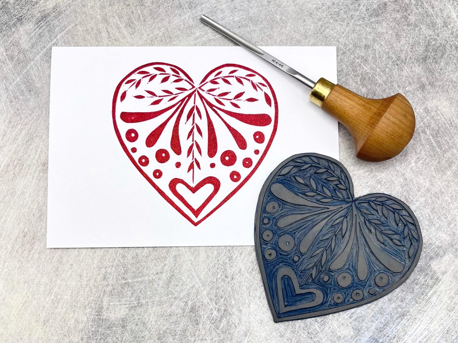 Red heart card from the set of three white cards with original lino printed heart designs. Laid on a textured surface. Photo includes lino and cutting tool.