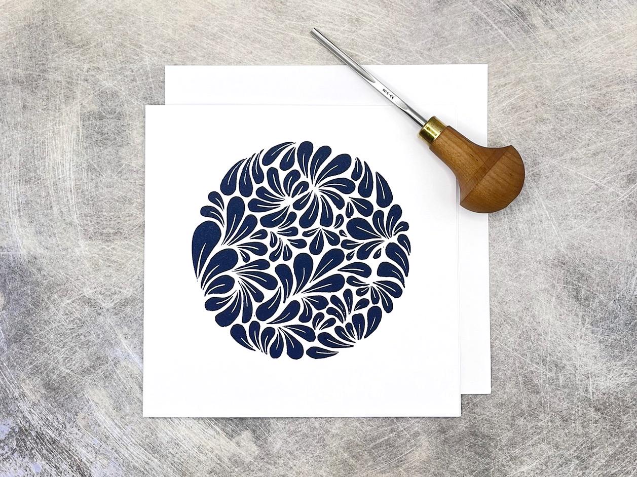 Navy blue original lino print, mussel shell pattern on a white card laid on a textured surface. Photo includes lino cutting tool used.