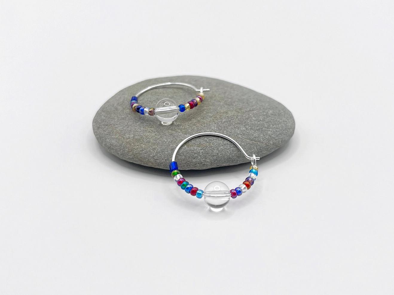 Rainbow seed bead and crystal hoop earrings laid on a grey pebble against a white background.