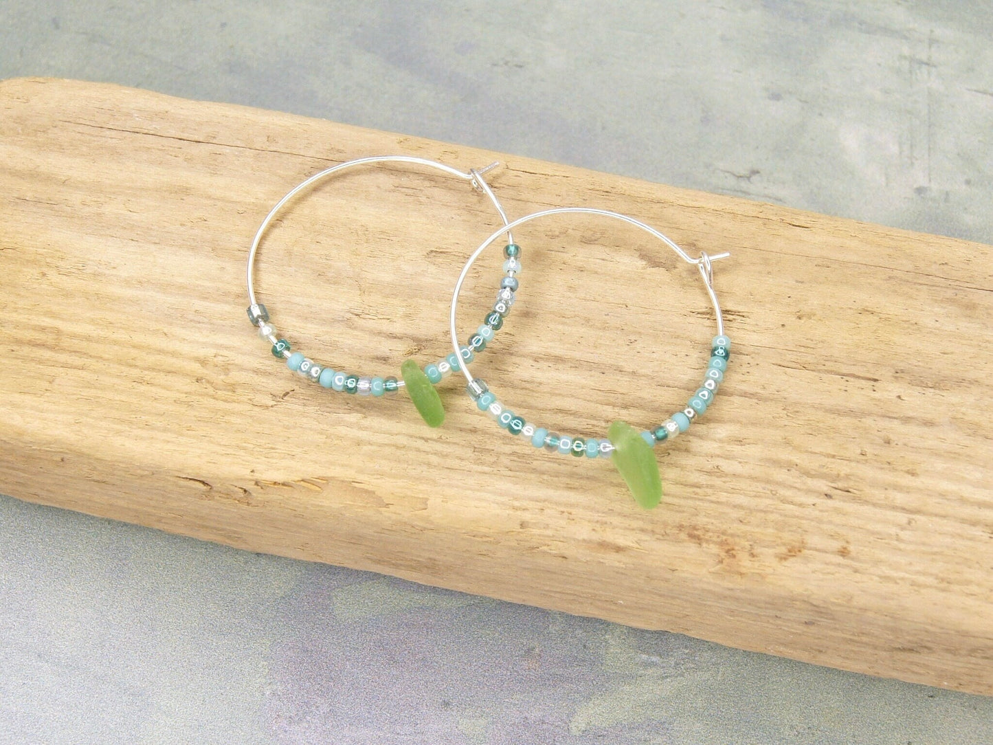Turquoise seed bead hoop earrings with green sea glass laid on a driftwood.