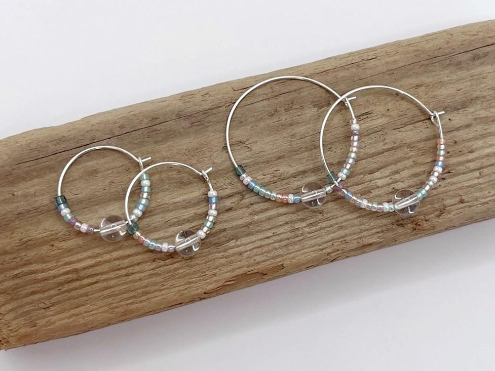 Two pairs of pastel seed bead and clear crystal hoop earrings laid on driftwood against a white surface
