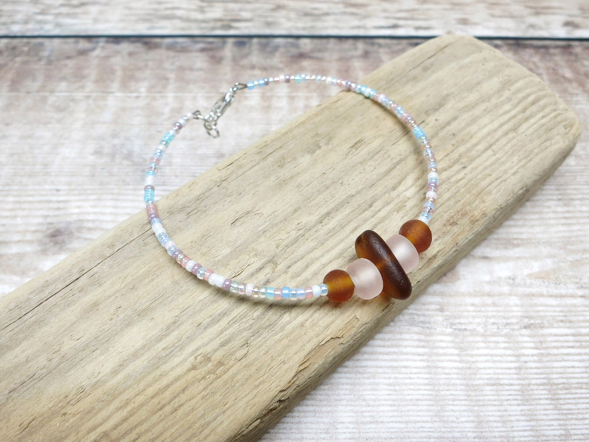 Bracelet with pastel seed beads and burnt orange sea glass laid on driftwood against a textured background.