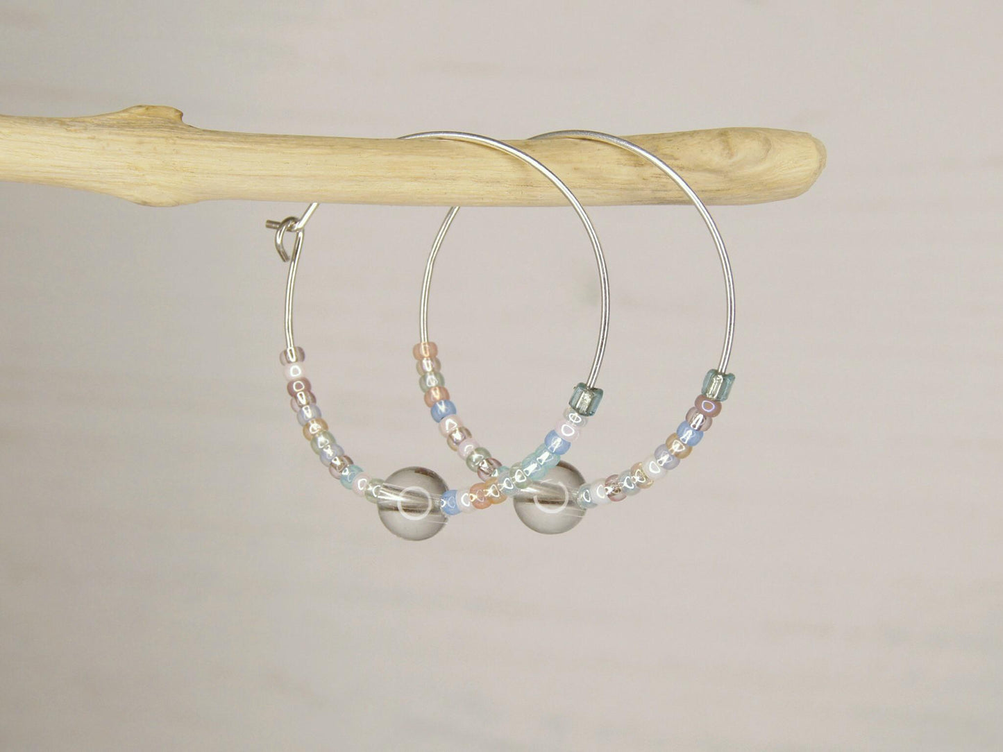 Pair of pastel seed bead and clear crystal hoop earrings hanging on driftwood against a pale background