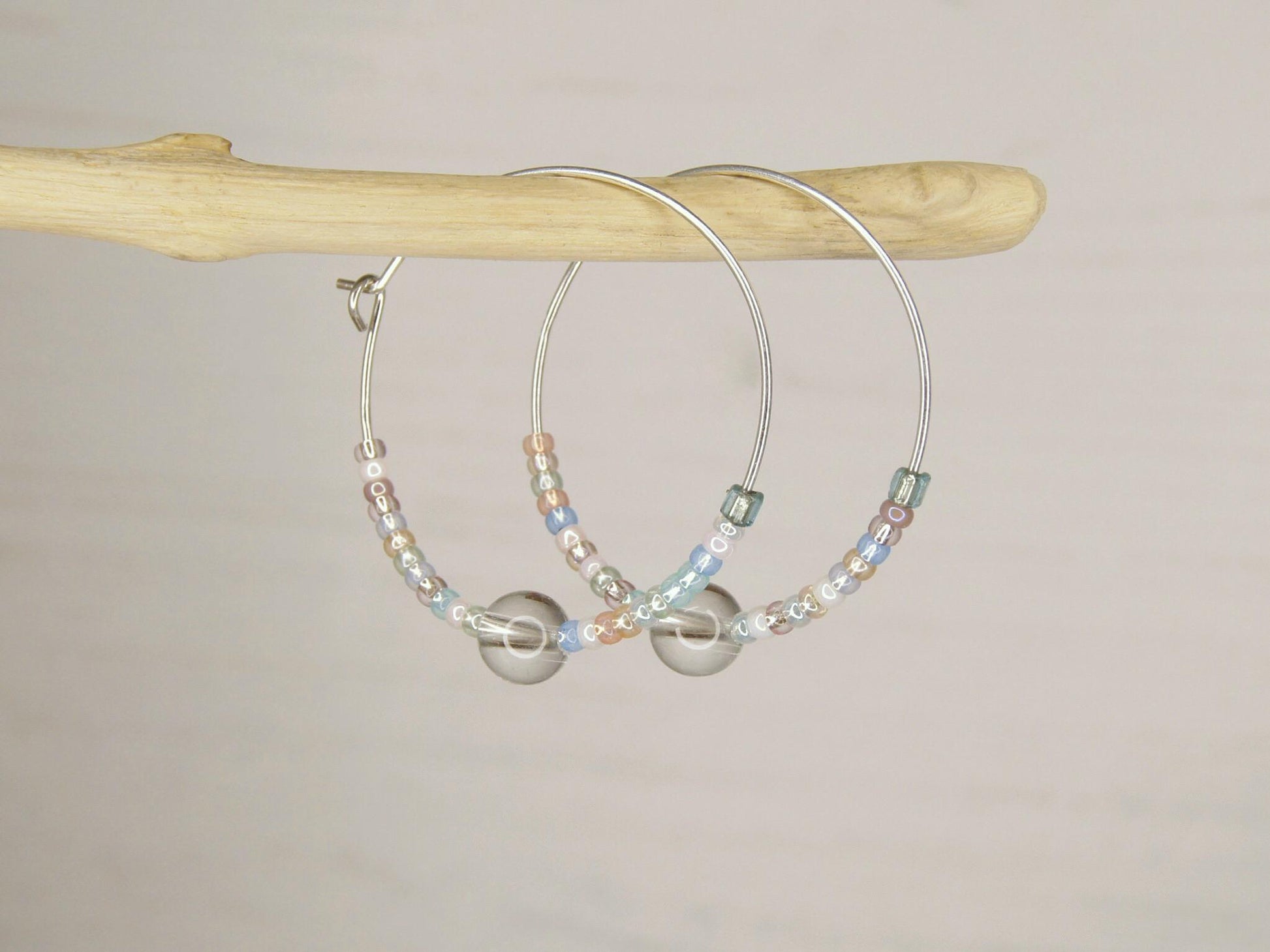 Pair of pastel seed bead and clear crystal hoop earrings hanging on driftwood against a pale background