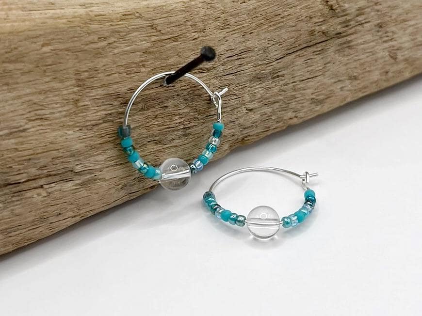 Silver hoop earrings with turquoise seed beads and crystal hanging on driftwood against a white background