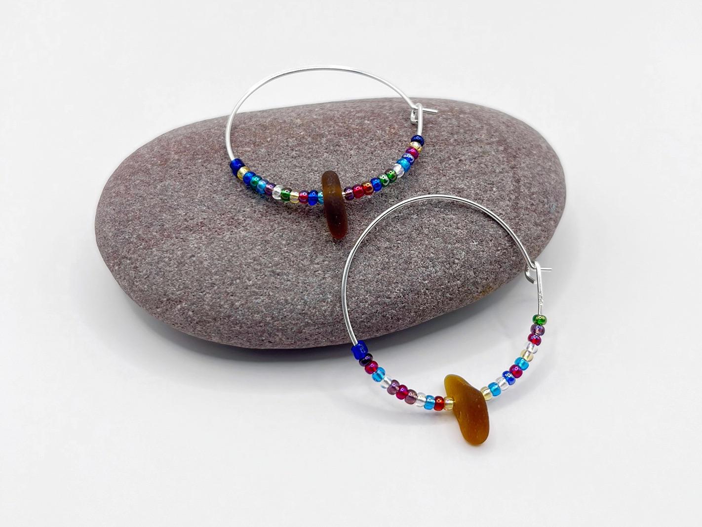 Rainbow seed bead and burnt orange sea glass hoop earrings laid on a purple pebble against a white background