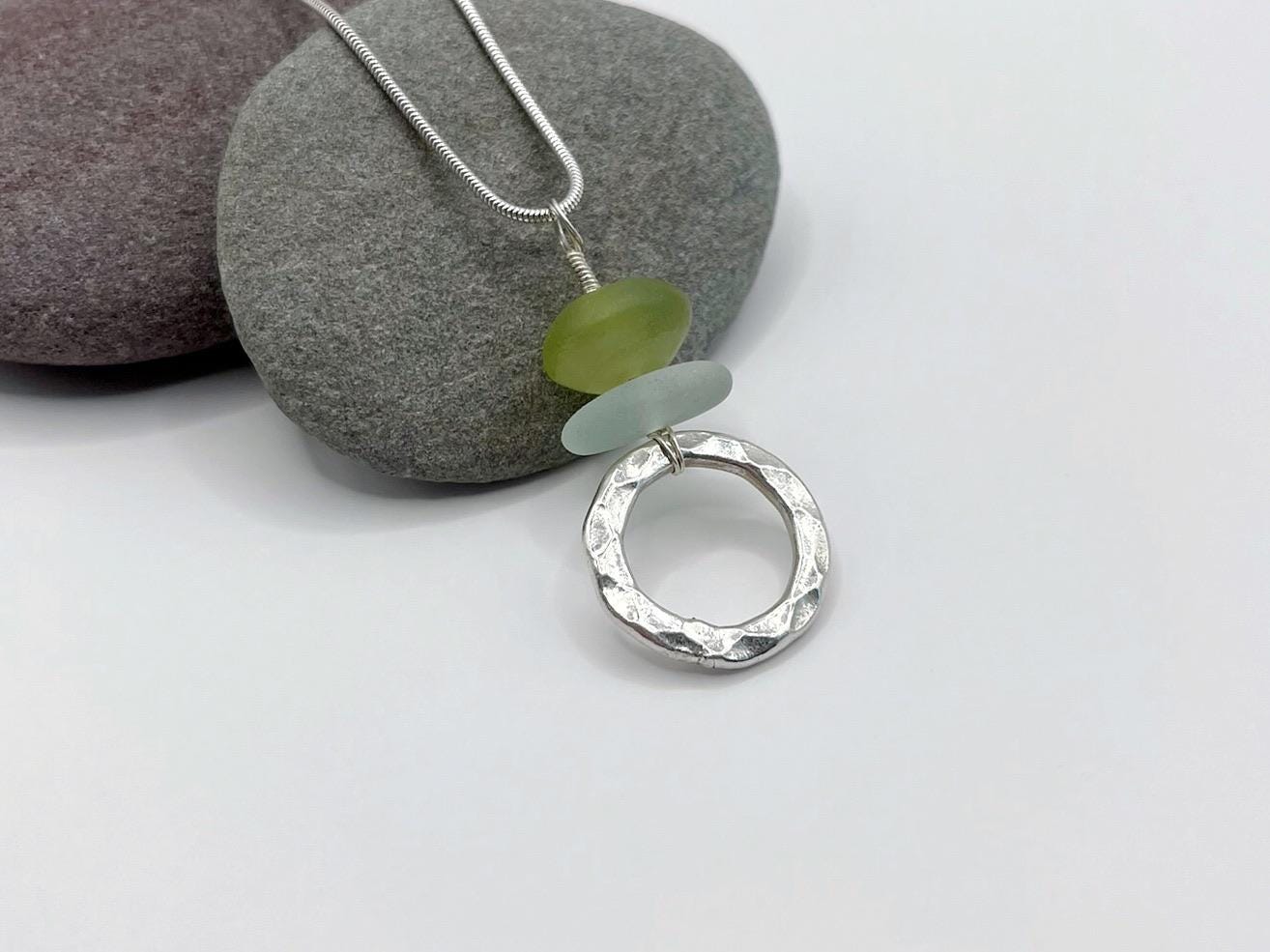 Silver necklace with a circular silver pendant, green jade and sea glass laid  on a grey pebble