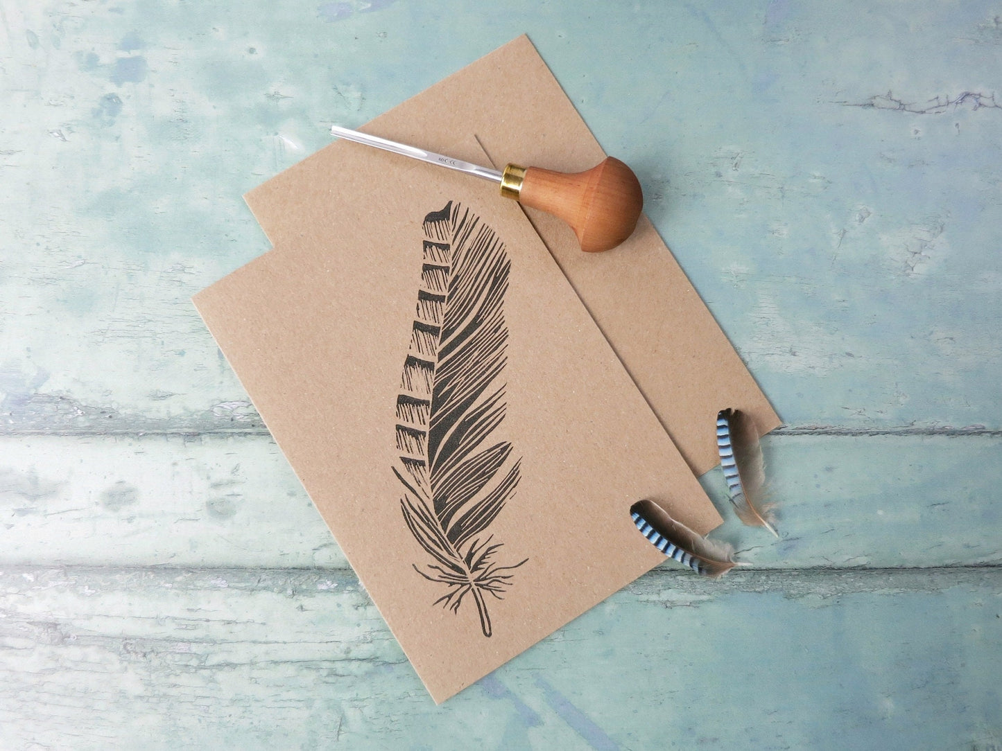 Kraft paper card with original Jay feather lino print laid on a textured surface. Photo includes lino cutting tool and two jay feathers