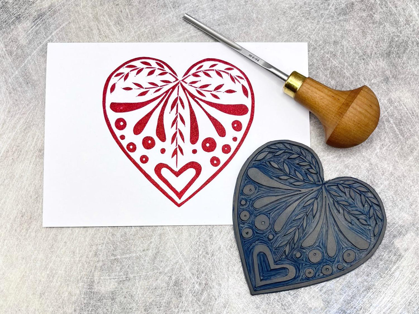White card with an original lino printed red heart design laid on a textured surface. Photo includes lino and cutting tool.