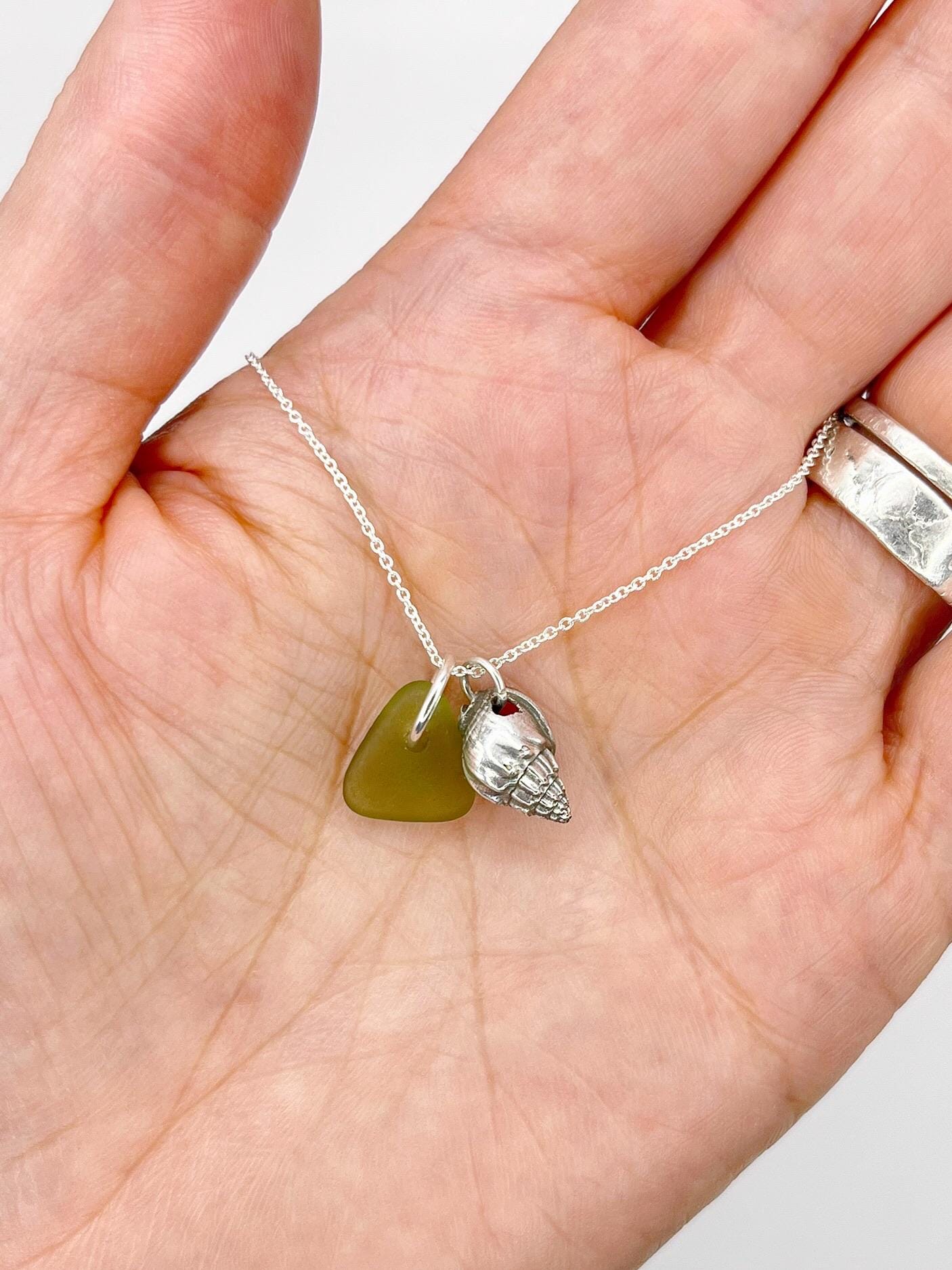 Hand holding a necklace with green sea glass and a silver shell charm against a white background