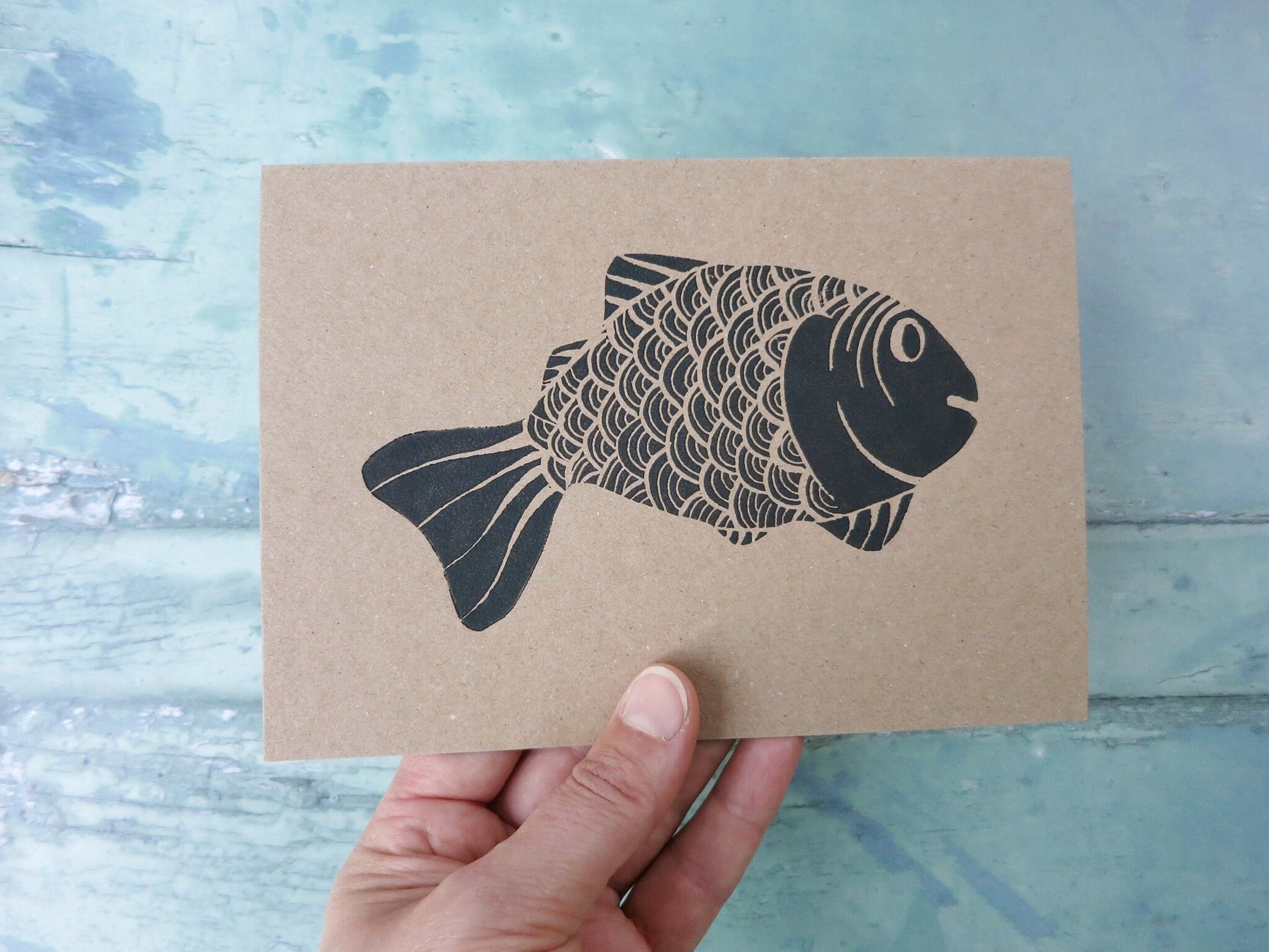 hand holding Kraft paper card with original lino printed fish against a textured surface. 