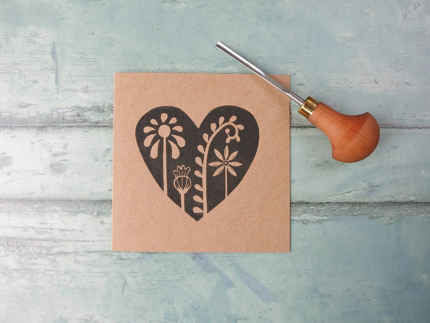 Kraft paper card with an original lino printed floral heart design laid on  a textured surface. Photo includes the lino cutting tool used.