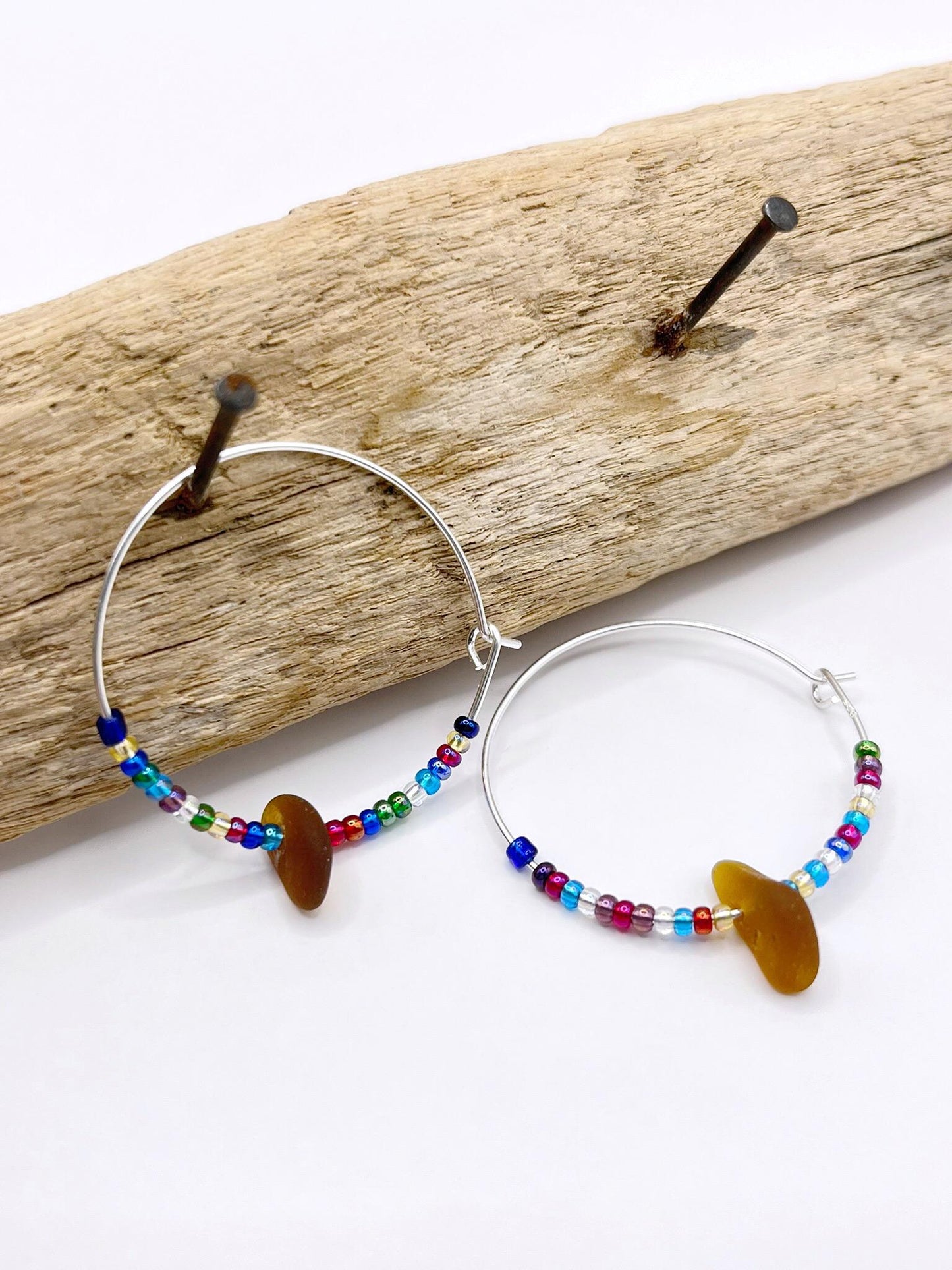 Rainbow seed bead and burnt orange sea glass hoop earrings hanging on driftwood against a white background