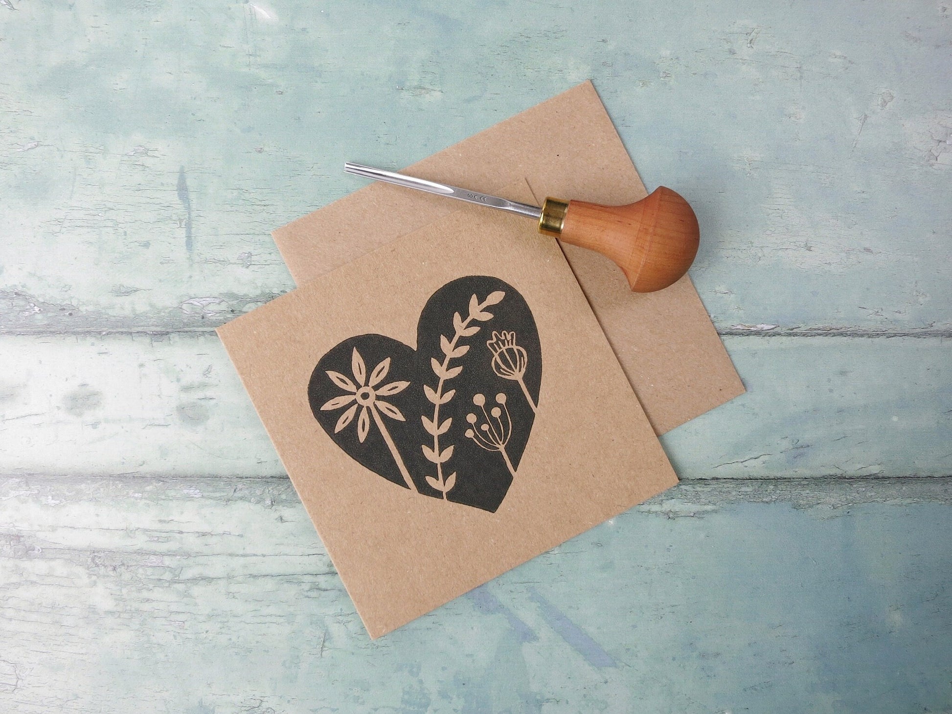 Kraft paper card with an original black lino printed heart design featuring floral elements laid on a textured surface. Photo includes lino cutting tool