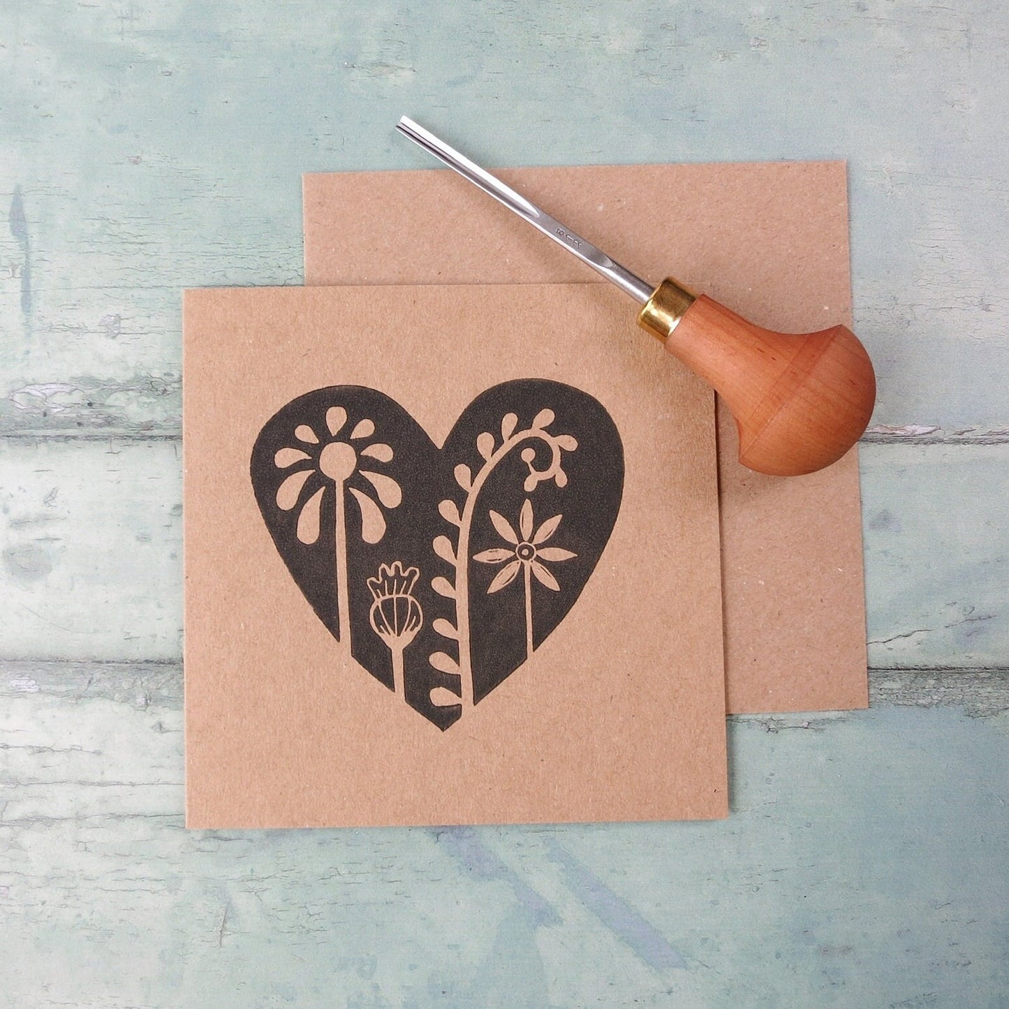 Kraft paper card with an original lino printed floral heart design laid on  a textured surface. Photo includes the lino cutting tool used.