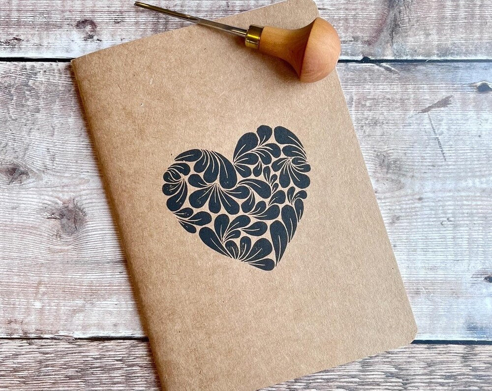 A5 Kraft notebook with original heart lino print on cover. Photo includes lino cutting tool used to make it. Laid on a textured background