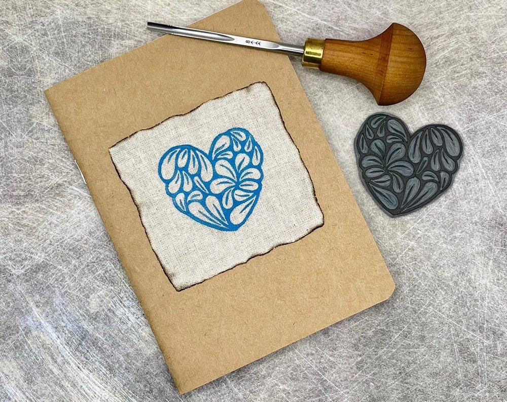 Close up of A6 Kraft sketchbook with original turquoise lino print on calico fabric on front. Photo includes lino and cutting tool used to make it.